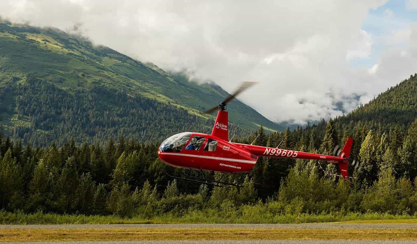 A red helicopter lifting off from a grassy field beside forest